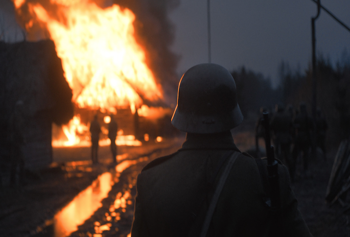 Eine Gruppe Soldaten und ein Soldat in Nahaufnahme von hinten in einer grauen Nacht stehen vor einem lichterloh brennenden Haus. Das Feuer reflektiert sich in den Pfützen