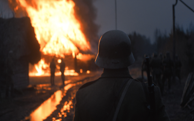 Eine Gruppe Soldaten und ein Soldat in Nahaufnahme von hinten in einer grauen Nacht stehen vor einem lichterloh brennenden Haus. Das Feuer reflektiert sich in den Pfützen